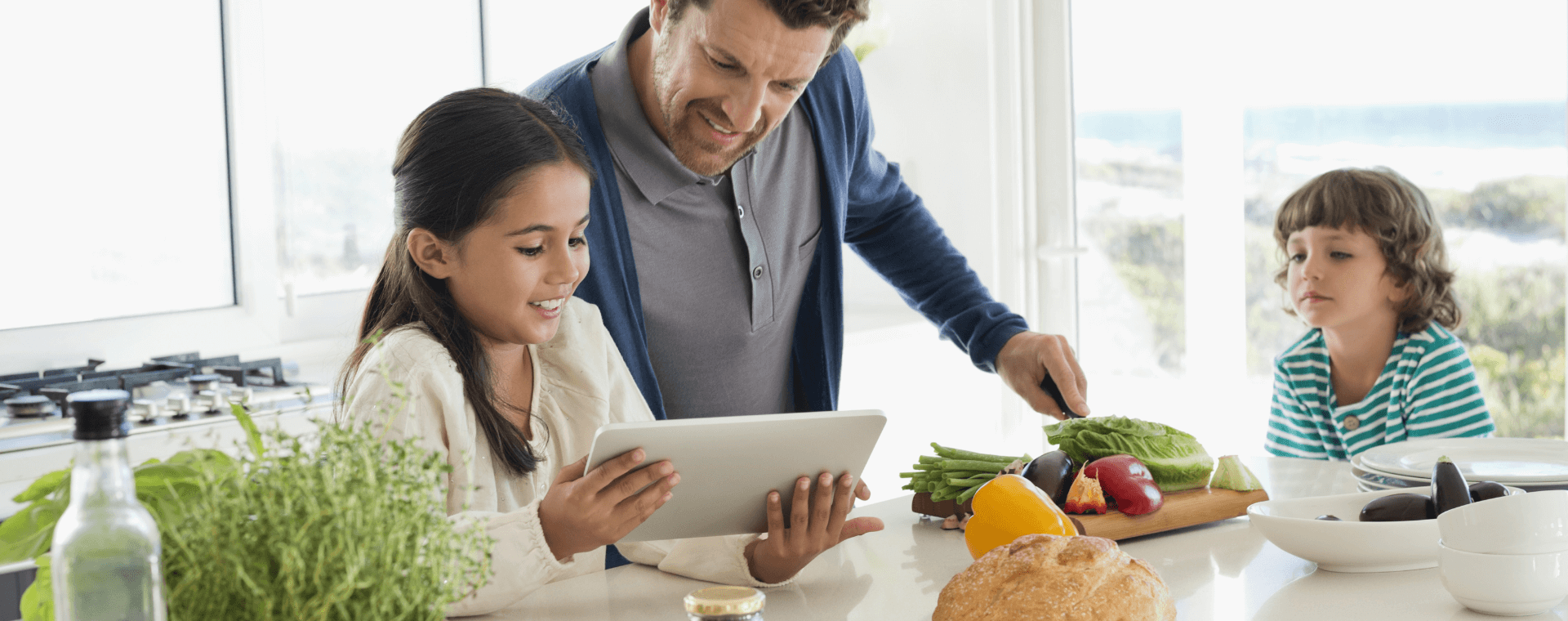 Father with his kids at kitchen counter reading a recipe off of a tablet.